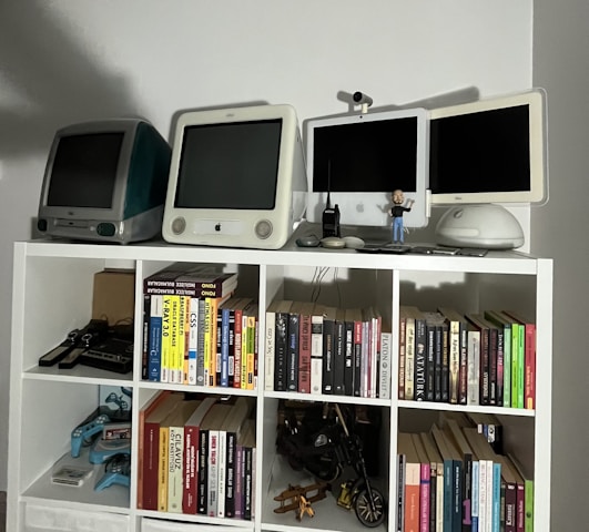 A collection of vintage computers and monitors displayed on a white shelving unit, accompanied by a variety of books neatly organized on the shelves. The top shelf features retro Apple computers and gadgets, including a webcam and a small figure. Below, books of different genres and topics line the shelves, with a few gaming controllers and a model motorcycle in the lower compartments.