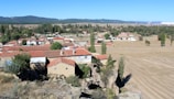 A rural landscape features a small village with houses topped with red-tiled roofs. The surrounding area consists of expansive fields and scattered trees, with a distant backdrop of hills under a clear blue sky.