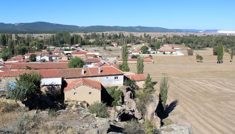 A rural landscape features a small village with houses topped with red-tiled roofs. The surrounding area consists of expansive fields and scattered trees, with a distant backdrop of hills under a clear blue sky.