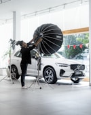 A person is adjusting a large studio light in front of a white SUV inside a showroom. The vehicle is positioned near large glass windows, and there are red triangular flags visible outside. The environment is bright and modern, with a sleek and minimal aesthetic.