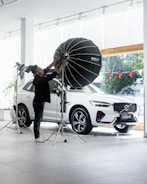 A person is adjusting a large studio light in front of a white SUV inside a showroom. The vehicle is positioned near large glass windows, and there are red triangular flags visible outside. The environment is bright and modern, with a sleek and minimal aesthetic.