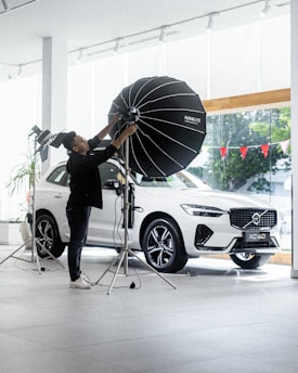 A person is adjusting a large studio light in front of a white SUV inside a showroom. The vehicle is positioned near large glass windows, and there are red triangular flags visible outside. The environment is bright and modern, with a sleek and minimal aesthetic.