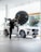 A person is adjusting a large studio light in front of a white SUV inside a showroom. The vehicle is positioned near large glass windows, and there are red triangular flags visible outside. The environment is bright and modern, with a sleek and minimal aesthetic.