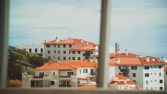 A cluster of white residential buildings with distinct red-orange roof tiles, captured from a window view. The architecture features multiple floors, balconies, and a few visible trees or shrubs surrounding the buildings. The scene appears bright with a clear sky in the background.