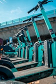 A row of teal-colored electric scooters lined up in an outdoor setting under a clear sky. The scooters are neatly arranged, and a concrete overpass is visible in the background, creating an urban environment.