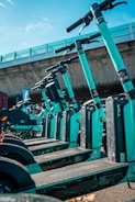 A row of teal-colored electric scooters lined up in an outdoor setting under a clear sky. The scooters are neatly arranged, and a concrete overpass is visible in the background, creating an urban environment.
