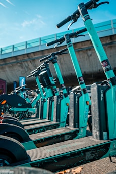 A row of teal-colored electric scooters lined up in an outdoor setting under a clear sky. The scooters are neatly arranged, and a concrete overpass is visible in the background, creating an urban environment.