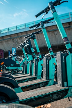 A row of teal-colored electric scooters lined up in an outdoor setting under a clear sky. The scooters are neatly arranged, and a concrete overpass is visible in the background, creating an urban environment.