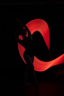 A silhouetted dancer poses gracefully against a backdrop of dynamic red light trails that form an abstract pattern, creating a captivating contrast with the dark background.