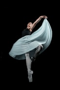 A graceful ballet dancer performing in an elegant pose, with one arm extended and the other holding a flowing, light-colored skirt. The dancer stands on pointed toes against a black background, highlighting the dynamic movement of the fabric.