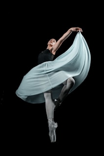 A graceful ballet dancer performing in an elegant pose, with one arm extended and the other holding a flowing, light-colored skirt. The dancer stands on pointed toes against a black background, highlighting the dynamic movement of the fabric.