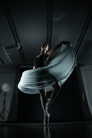 A ballerina gracefully balances on pointe, her elegant pose emphasized by a flowing skirt that catches the light. The dimly lit studio showcases dramatic shadows, enhancing the sense of movement and poise.
