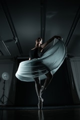 A ballerina gracefully balances on pointe, her elegant pose emphasized by a flowing skirt that catches the light. The dimly lit studio showcases dramatic shadows, enhancing the sense of movement and poise.