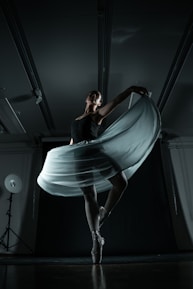 A ballerina gracefully balances on pointe, her elegant pose emphasized by a flowing skirt that catches the light. The dimly lit studio showcases dramatic shadows, enhancing the sense of movement and poise.