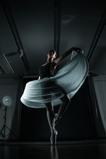 A ballerina gracefully balances on pointe, her elegant pose emphasized by a flowing skirt that catches the light. The dimly lit studio showcases dramatic shadows, enhancing the sense of movement and poise.