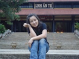 A person is sitting on stone steps leading up to a traditional temple with a sign that reads 'LINH ẢN TỰ'. The person is smiling and resting their head on their hand. The temple is adorned with a large roof and surrounded by greenery.