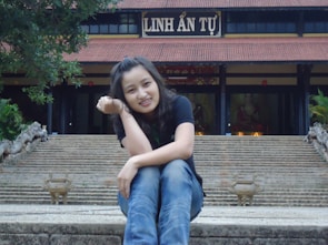 A person is sitting on stone steps leading up to a traditional temple with a sign that reads 'LINH ẢN TỰ'. The person is smiling and resting their head on their hand. The temple is adorned with a large roof and surrounded by greenery.