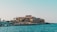 A seaside view of a large building labeled as a casino, situated on a rocky shore. The building is surrounded by palm trees and other smaller structures. The calm sea water reflects the sky, and there are several closed umbrellas lined along the waterfront.