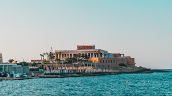 A seaside view of a large building labeled as a casino, situated on a rocky shore. The building is surrounded by palm trees and other smaller structures. The calm sea water reflects the sky, and there are several closed umbrellas lined along the waterfront.