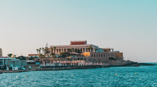 A seaside view of a large building labeled as a casino, situated on a rocky shore. The building is surrounded by palm trees and other smaller structures. The calm sea water reflects the sky, and there are several closed umbrellas lined along the waterfront.