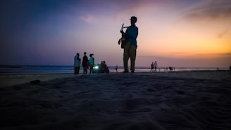 A silhouette of a person playing guitar on a beach during sunset. In the background, several other people are gathered in small groups, some near the edge of the water. The sky is transitioning from blue to shades of orange, pink, and purple.