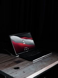 A laptop sits on a wooden desk in a dimly lit environment. The screen displays a login page with red, white, and gray graphics. The laptop is a MacBook, identifiable by the logo on the screen. A power cable is connected to the laptop.