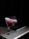 A laptop sits on a wooden desk in a dimly lit environment. The screen displays a login page with red, white, and gray graphics. The laptop is a MacBook, identifiable by the logo on the screen. A power cable is connected to the laptop.