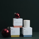 Several boxed candles labeled with words such as 'Strength', 'Hope', and 'Peace' are arranged in a stacked formation against a dark background. A red Christmas ornament is placed on top of one of the boxes, adding a festive touch.