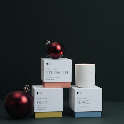 Several boxed candles labeled with words such as 'Strength', 'Hope', and 'Peace' are arranged in a stacked formation against a dark background. A red Christmas ornament is placed on top of one of the boxes, adding a festive touch.