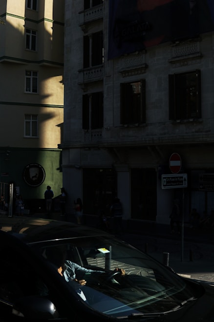 A street scene in an urban environment with a darkened, high-contrast lighting. Sunlight casts shadows on the facade of a building, highlighting its textures and windows. A car is partially visible in the lower part of the image, with a person inside possibly using a phone or device. Traffic signs and a few people are present on the sidewalk, blending into the shadows.