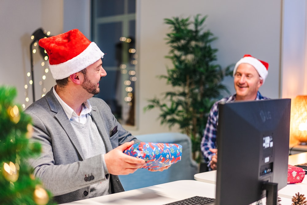 a man in a santa hat and a man in a suit sitting at a desk with a laptop
