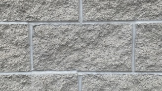 A close-up view of a wall constructed from rough gray concrete blocks arranged in a neat, grid-like pattern. The texture is coarse and slightly uneven, with visible grout lines separating each block.