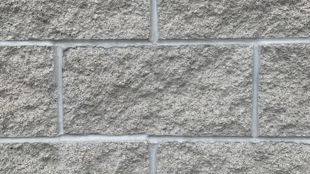 A close-up view of a wall constructed from rough gray concrete blocks arranged in a neat, grid-like pattern. The texture is coarse and slightly uneven, with visible grout lines separating each block.