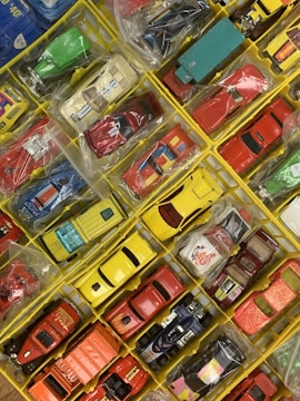 A collection of colorful toy cars organized in a grid of yellow compartments. Each car is individually placed in clear plastic bags, displaying a variety of designs, colors, and styles including sports cars, trucks, and vintage models. The arrangement is tidy and visually appealing.