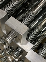 A close-up view of multiple metallic threaded rods aligned neatly in parallel. The threads on the rods are evenly spaced and the metallic surfaces are polished, reflecting light and creating shiny highlights.