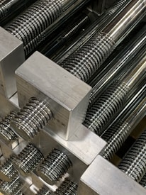 A close-up view of multiple metallic threaded rods aligned neatly in parallel. The threads on the rods are evenly spaced and the metallic surfaces are polished, reflecting light and creating shiny highlights.