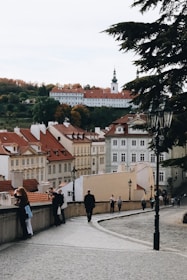 A picturesque European cityscape featuring rows of historic buildings with red rooftops. The scene includes a church with a tall spire in the background and a hill with autumn-colored trees. In the foreground, a cobblestone path lined with street lamps is occupied by several people walking or taking photos.