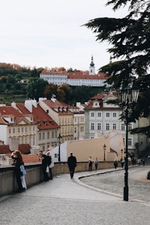 A picturesque European cityscape featuring rows of historic buildings with red rooftops. The scene includes a church with a tall spire in the background and a hill with autumn-colored trees. In the foreground, a cobblestone path lined with street lamps is occupied by several people walking or taking photos.