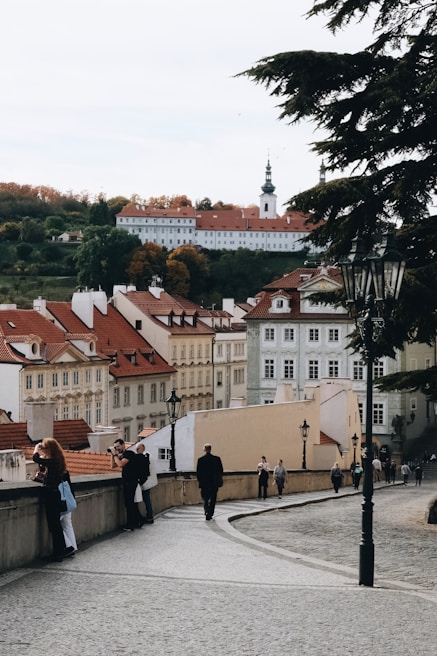 A picturesque European cityscape featuring rows of historic buildings with red rooftops. The scene includes a church with a tall spire in the background and a hill with autumn-colored trees. In the foreground, a cobblestone path lined with street lamps is occupied by several people walking or taking photos.