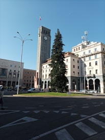 An urban square with a tall clock tower, flying the Italian flag at the top. Surrounding buildings feature classical architecture with arched windows and ornate detailing. A large evergreen tree is centrally positioned in the square, and several cars and pedestrians are visible nearby.