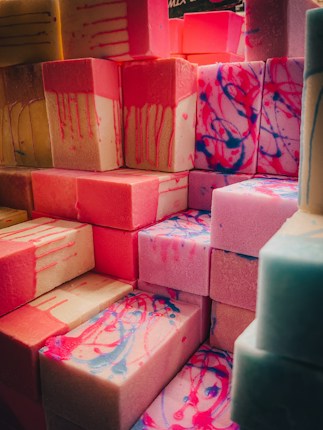 A stack of colorful soap bars with various patterns and shades, featuring drips and swirls. The dominant colors include pink, blue, and beige, creating a visually appealing and artistic arrangement.