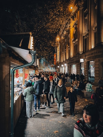 A bustling street market scene at night, with people walking along a cobblestone path lined with illuminated wooden stalls. The atmosphere is lively as individuals browse and engage with vendors. Decorative lights and the warm glow from the stalls create a cozy ambiance. The architecture of the nearby building is grand, with tall columns and lit facades adding to the city's festive vibe.