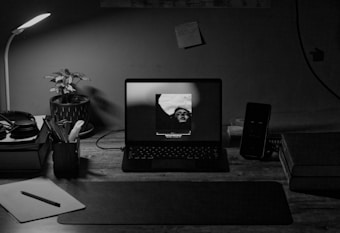 A desk setup featuring an open laptop displaying a black-and-white image of a person. Surrounding the laptop are various items including a desk lamp, a potted plant, headphones, notebooks, a smartphone, and a lined notepad with a pen. The lighting is dim, coming primarily from the lamp and the laptop screen, creating a cozy and focused atmosphere.