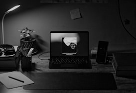 A desk setup featuring an open laptop displaying a black-and-white image of a person. Surrounding the laptop are various items including a desk lamp, a potted plant, headphones, notebooks, a smartphone, and a lined notepad with a pen. The lighting is dim, coming primarily from the lamp and the laptop screen, creating a cozy and focused atmosphere.
