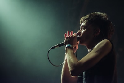 A person is singing into a microphone with a spotlight shining from behind. The person is wearing a sleeveless top and appears to be in a dark, possibly concert, setting.