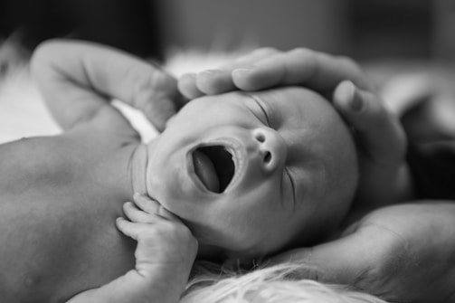 A newborn baby rests while yawning, with a gentle hand placed on the head. The baby's eyes are closed, and the hand provides a sense of comfort and protection.