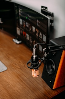 A workspace setup featuring a wooden desk with a mounted microphone. The microphone is positioned above the desk, indicating a recording setup. In the background, there is a large monitor showing a digital music interface, and a speaker with a wood finish is visible beside it.