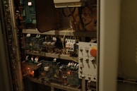 A complex electrical control panel with numerous wires, switches, and circuit boards. A large red emergency stop button is prominently visible, along with various control switches and labels in Russian. The panel is housed inside a metal cabinet, with several electronic components emitting faint lights.