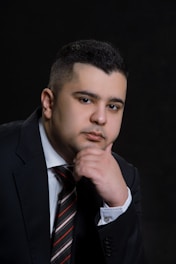 A person in a formal suit and tie, resting their chin on their hand, against a dark background. The individual is gazing forward with a contemplative expression, wearing a dark jacket, white shirt, and striped tie.
