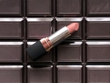 A pink lipstick rests on a background featuring a pattern resembling a chocolate bar. The lipstick has a sleek silver and black casing with a rose gold accent. The chocolate bar background has distinct, glossy square patterns.